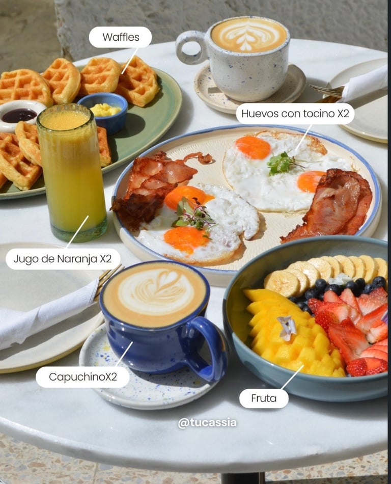 Breakfast spread with waffles, eggs, bacon toast, fresh fruit, orange juice, and cappuccinos with latte art on a white table