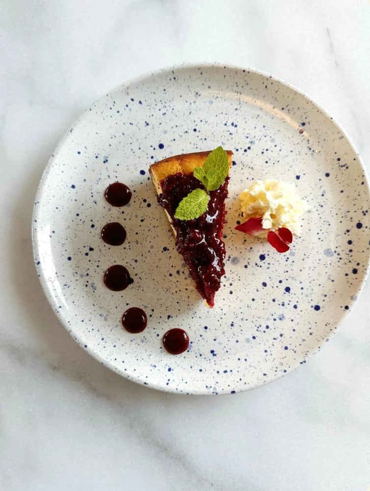 Elegant plated dessert on speckled white plate with dark berry tart slice, chocolate dots, mint leaf, and cream garnish